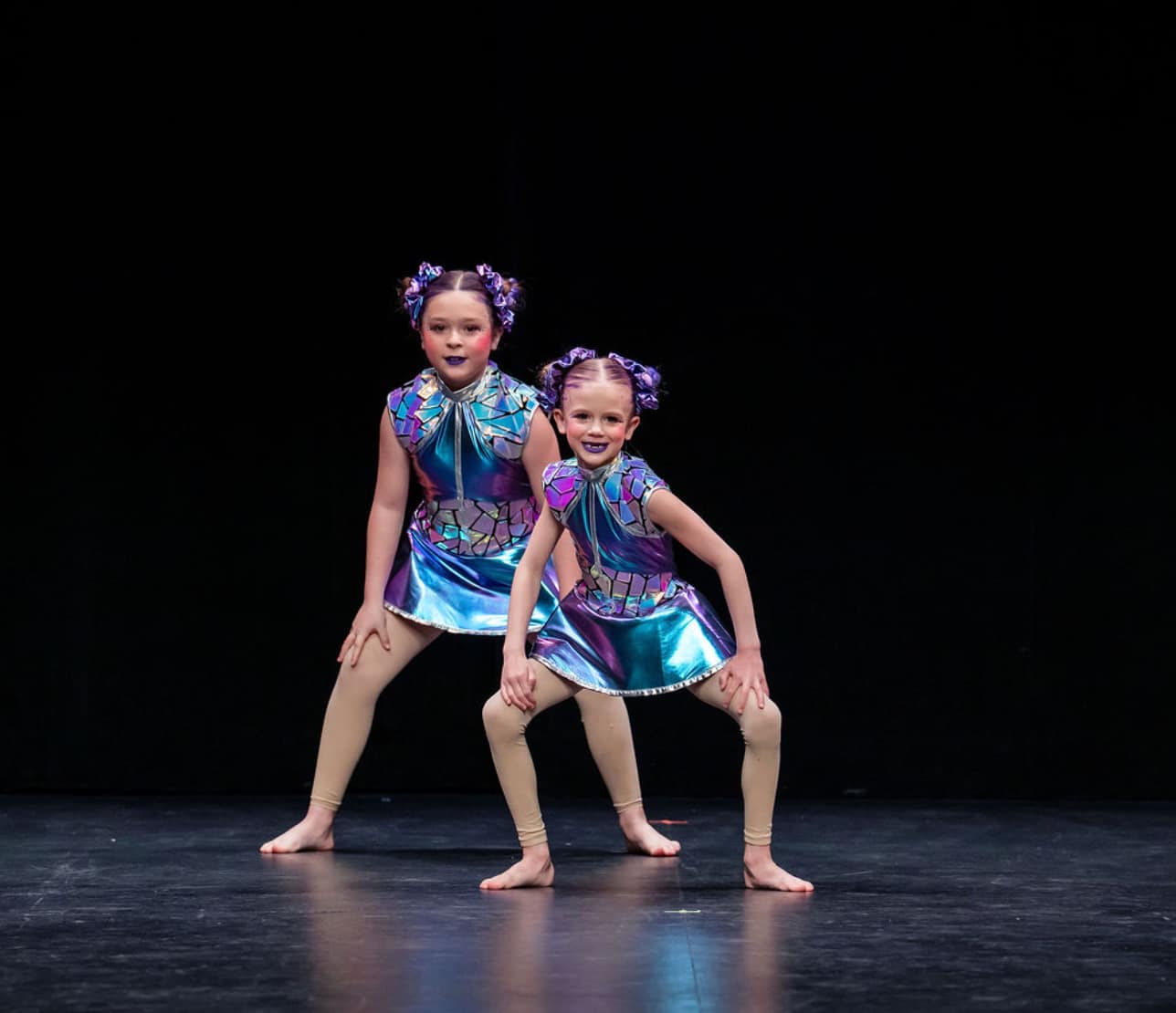 Two young dancers in metallic purple costumes performing a hip hop pose on stage